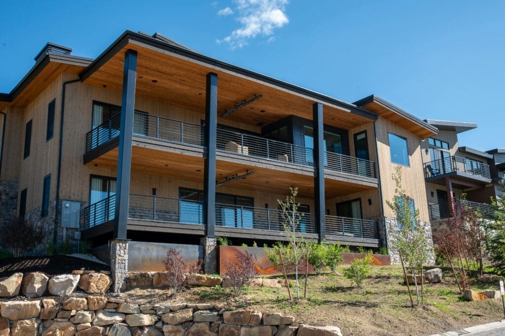 Modern two-story apartment building with wooden siding, large balconies, and black metal railings, surrounded by newly planted landscaping and stone retaining walls under a clear blue sky.